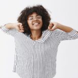 Waist-up shot of energetic cute african american female who slept well stretching hands and smiling happily after great nap being ready to work productively after good rest over gray background.